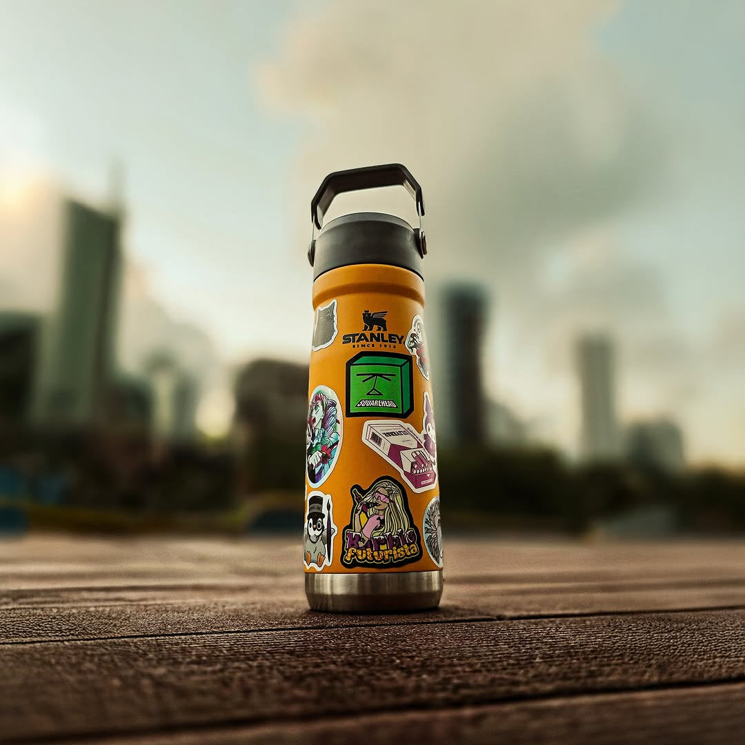 Garrafa térmica Stanley laranja com vários adesivos personalizados, sobre mesa de madeira e fundo urbano desfocado.