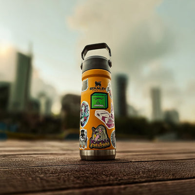 Garrafa térmica Stanley laranja com vários adesivos personalizados, sobre mesa de madeira e fundo urbano desfocado.